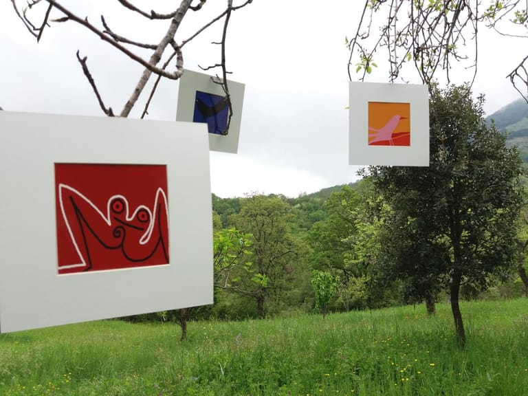 Tre opere in passe-partout bianco appese ai rami di alberi in un prato