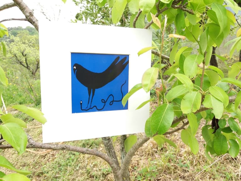 Opera con uccello nero su fondo blu, montata in un passe-partout bianco appeso tra i rami di un albero